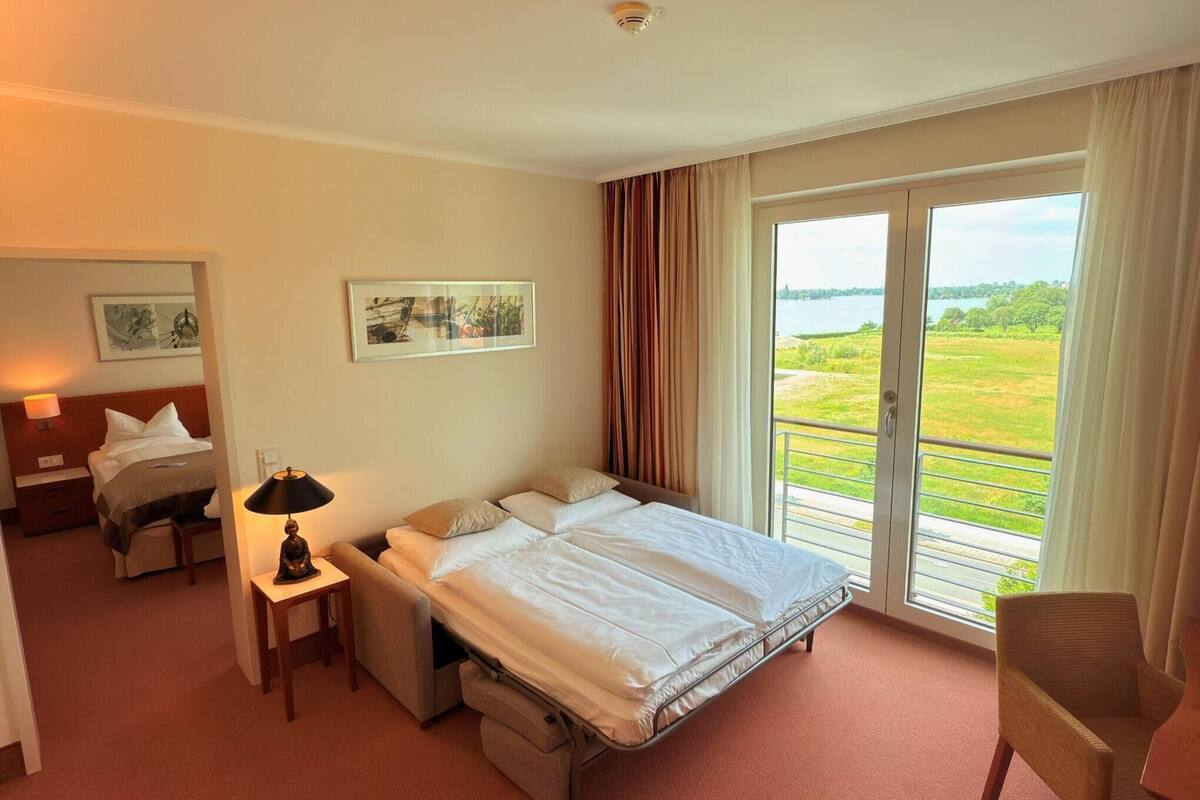 A hotel room with a bed, a desk, a chair, a lamp, and a large window with a view of a green landscape.