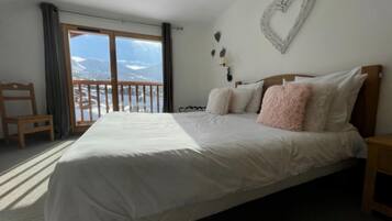 Een slaapkamer met een groot bed, een houten hoofdbord, een wanddecoratie in de vorm van een hart en uitzicht op bergen vanaf een balkon.