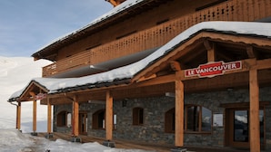 A wooden chalet with a stone facade, a balcony with snow-covered railings, and a sign reading "LE VANCOUVER".