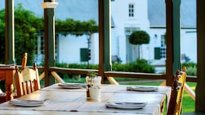 Une table à manger est dressée avec des assiettes et un verre d’eau, et offre une vue sur un bâtiment blanc et sur la verdure en arrière-plan.