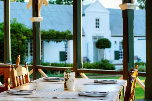 Una mesa lista con platos y un vaso de agua, con vista a un edificio blanco y vegetación al fondo.