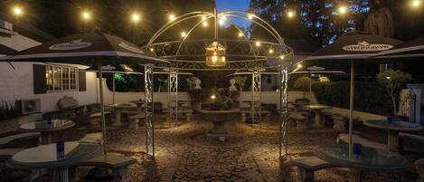 An outdoor patio area with stone tables and chairs, a central fountain, and string lights.