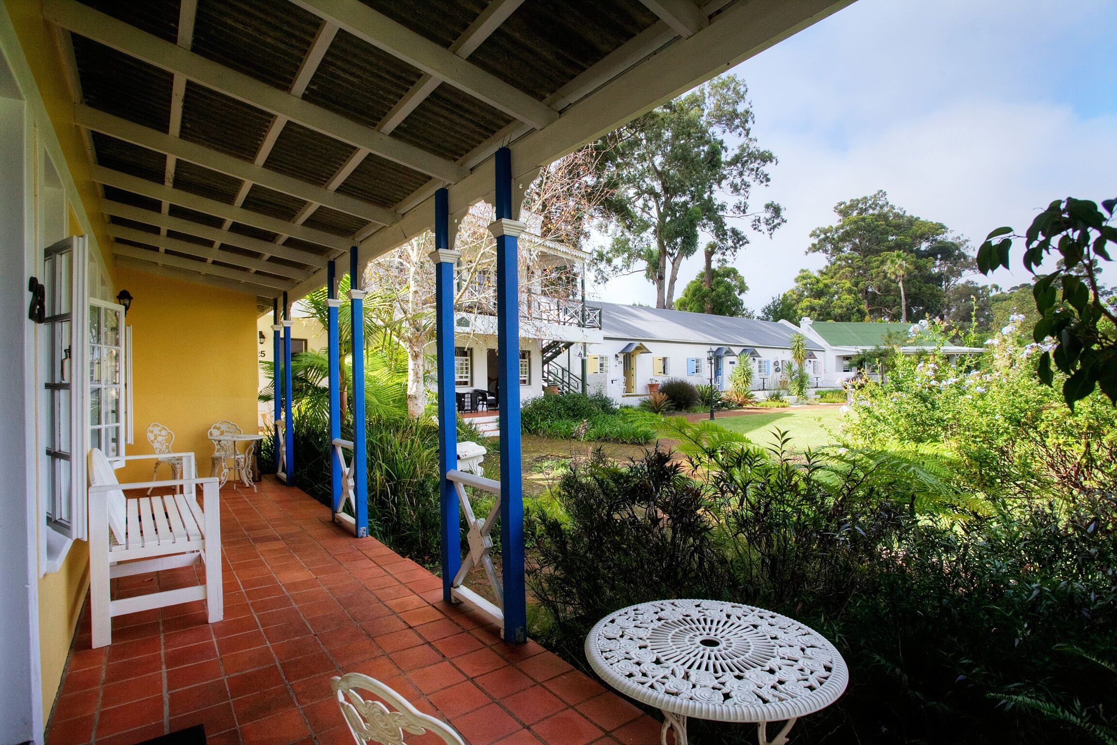 A covered patio with white furniture, a decorative table, and a view of a garden and house in the background.
