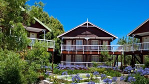 A two-story wooden cabin with a balcony, surrounded by lush greenery and flowering plants.