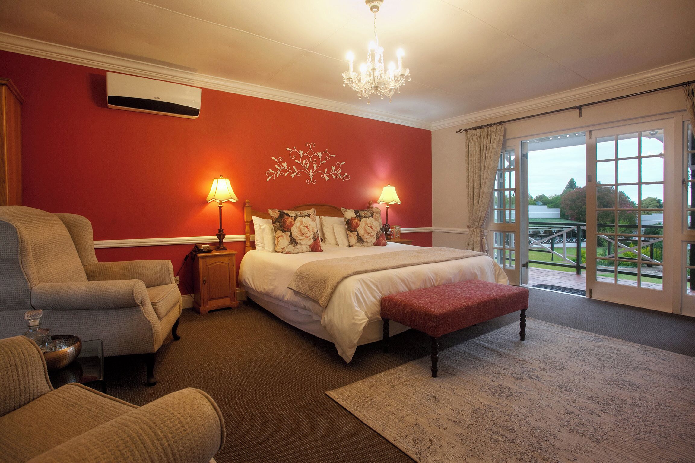 A bedroom with a large bed, two bedside lamps, a chandelier, a red wall, and a view through the glass doors.