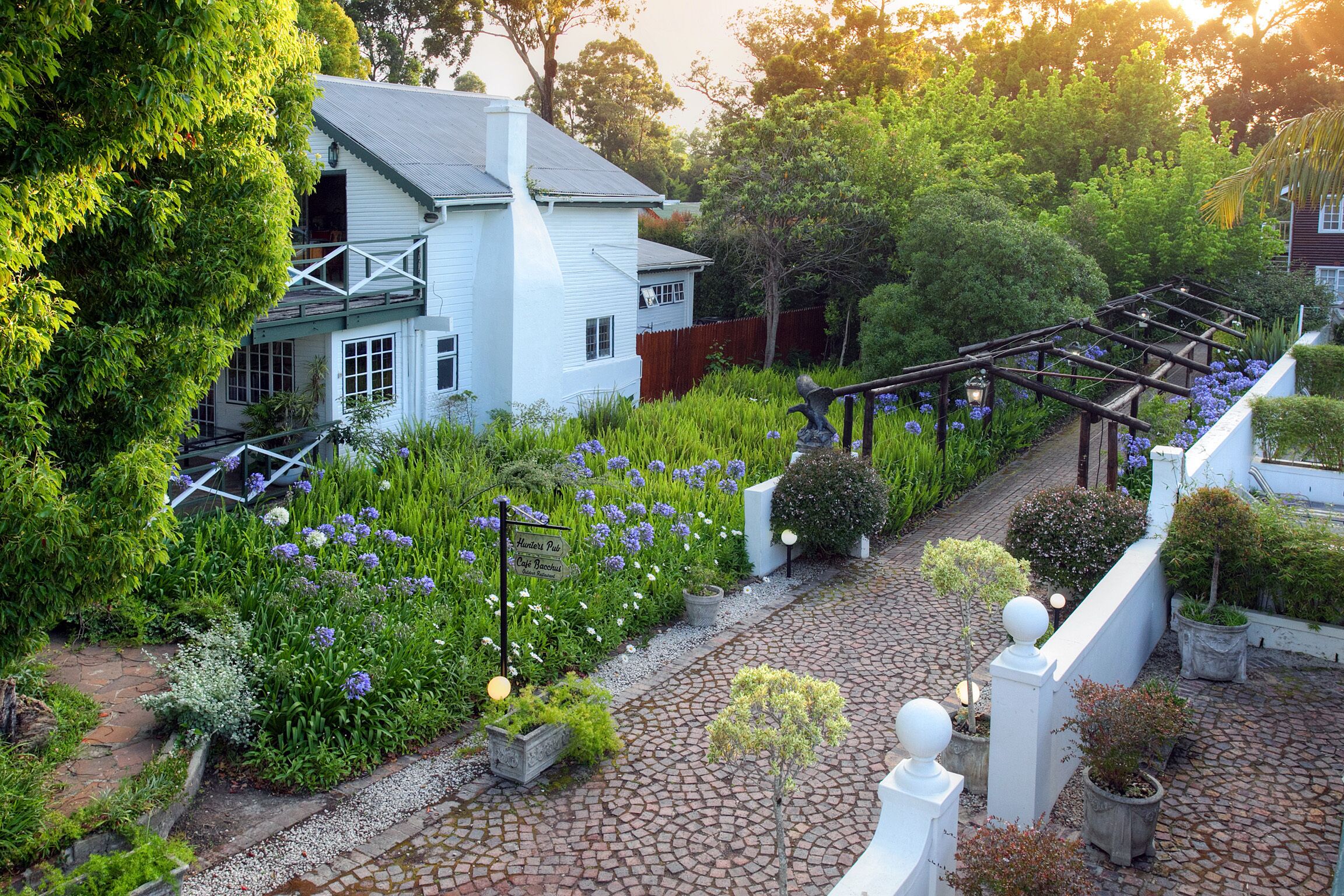 A white house with a balcony, surrounded by lush greenery and a garden with purple flowers.