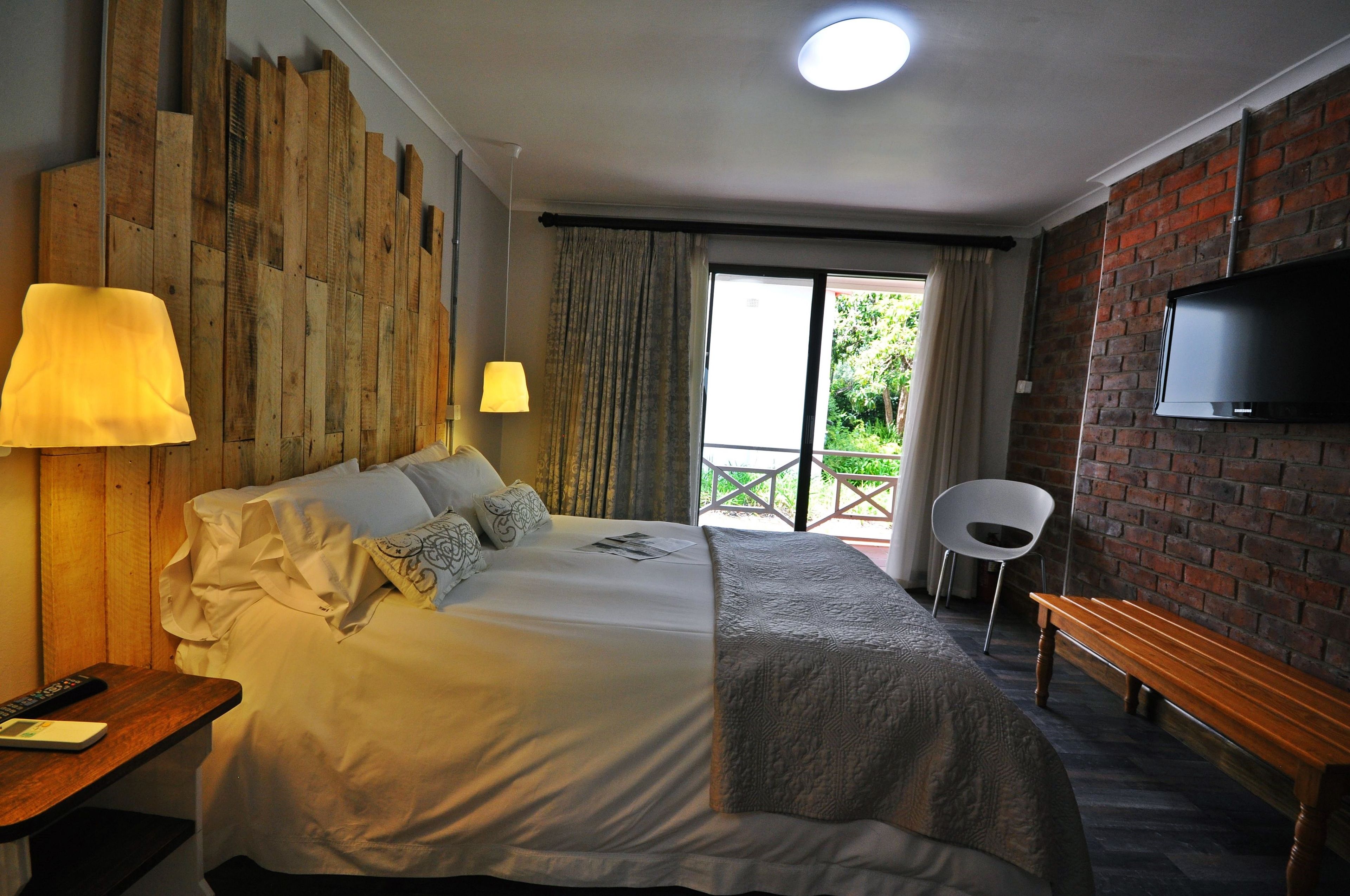 Een hotelkamer met een groot bed, twee bedlampen, een aan een bakstenen muur bevestigde televisie, een houten bankje en een balkon met een stoel.