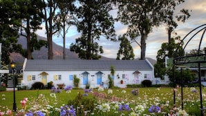 Un bâtiment blanc aux portes et fenêtres bleues, entouré d’un jardin où poussent différentes sortes de fleurs et d’arbres.
