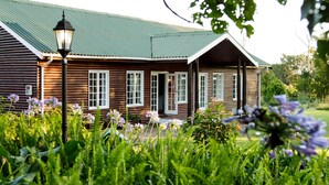 A wooden cabin with a green roof, surrounded by lush greenery and purple flowers.