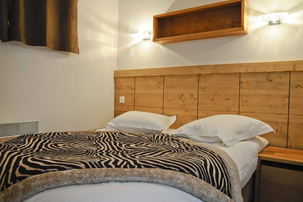 A bedroom with a wooden headboard, a zebra-patterned bedspread, white pillows, and a wooden shelf with wall-mounted lights.