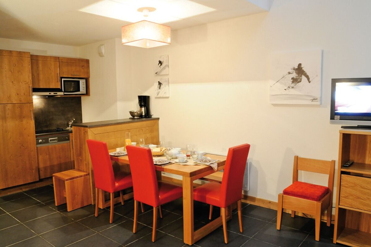 A compact kitchen with a microwave, stove, and coffee maker. There is a dining table with four red chairs and a small wooden stool. A television is mounted on the wall to the right.