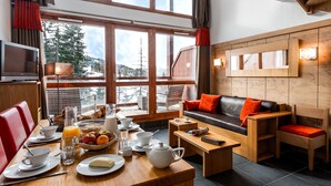 Una habitación acogedora con una mesa de comedor dispuesta para el desayuno, un sofá con cojines rojos y un gran ventanal con vistas a montañas nevadas.