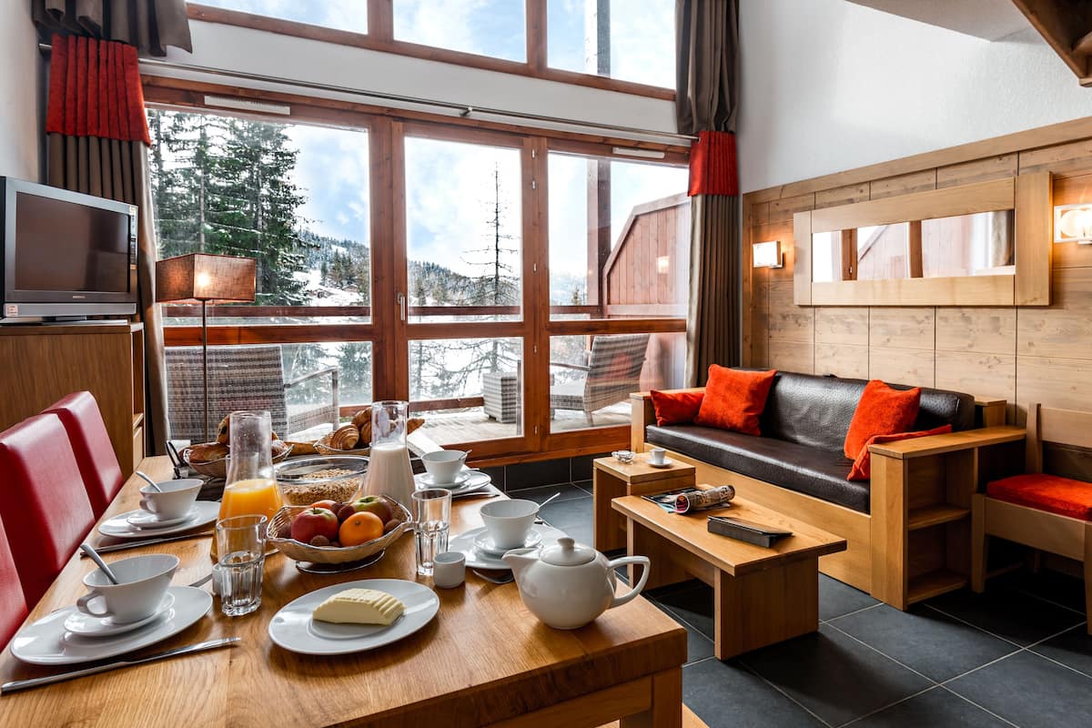 A cozy room with a dining table set for breakfast, a sofa with red cushions, and a large window offering a view of snowy mountains.