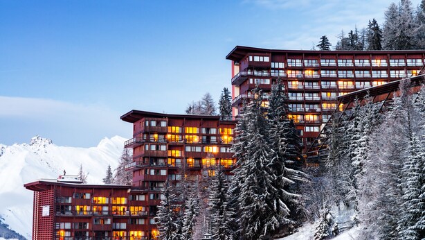 Un hotel de varios pisos con balcones y ventanas amplias, rodeado de árboles y montañas cubiertas de nieve.