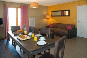 Uma área de refeições com mesa de madeira posta para o pequeno-almoço, incluindo croissants e sumo de laranja. Há um sofá com almofadas e uma lâmpada ao fundo.