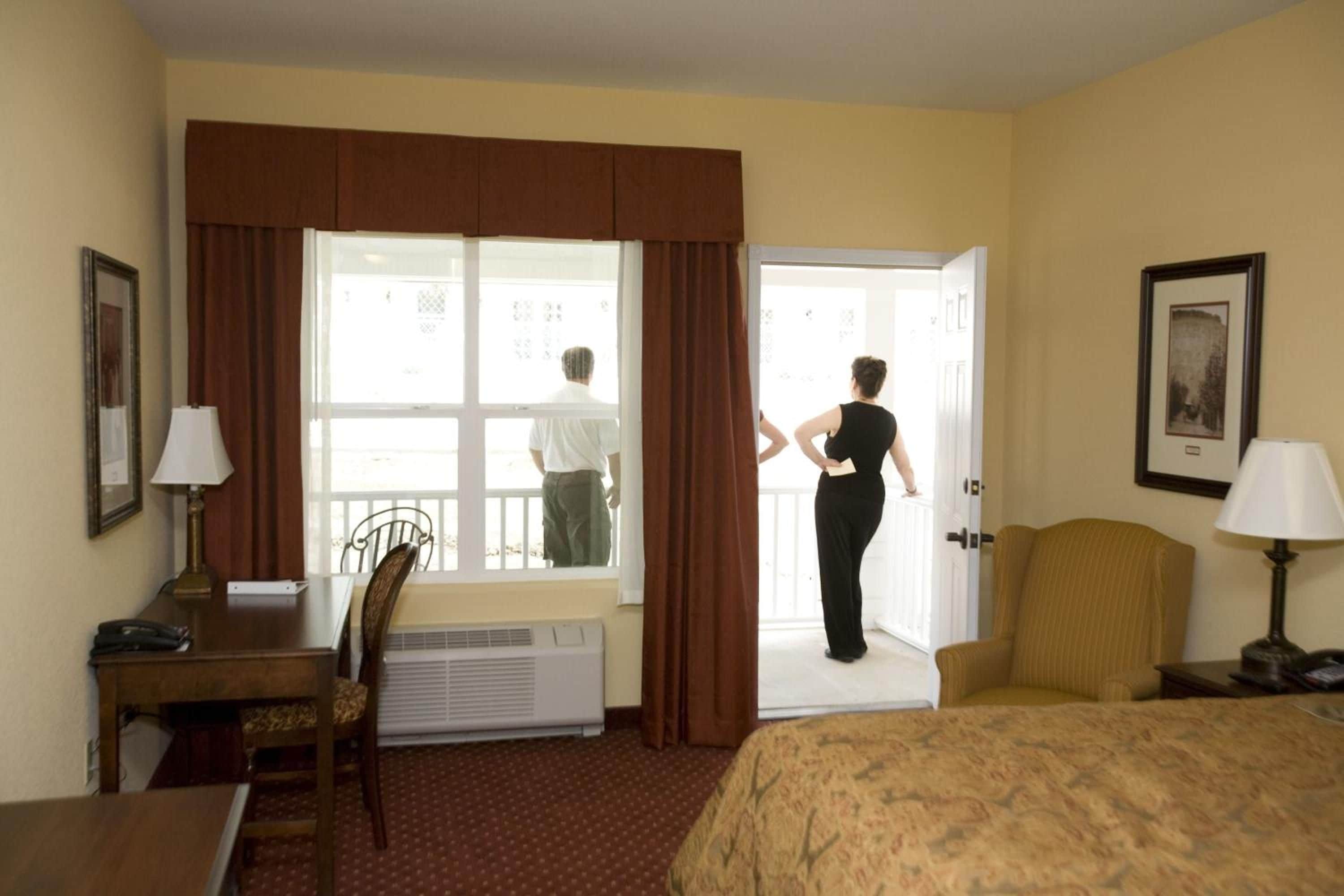 A hotel room with a bed, a desk, a chair, a window with curtains, and a door.