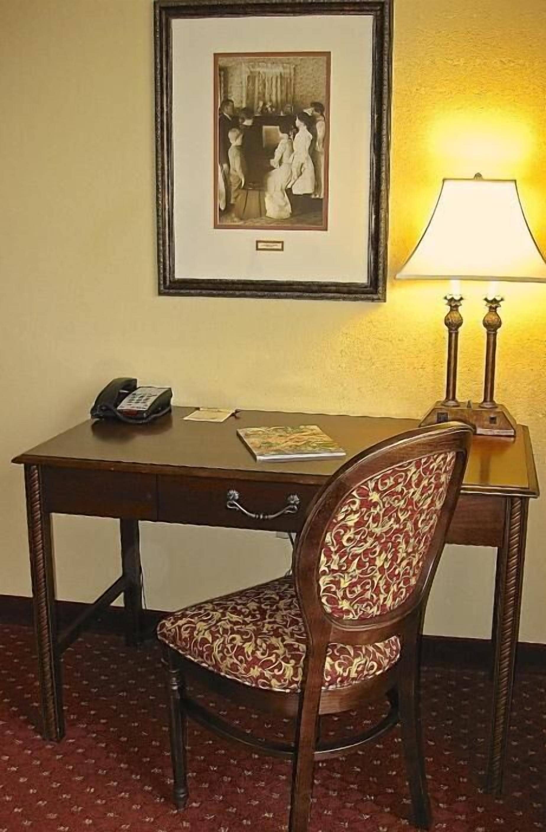 Ein Hotelzimmer mit einem Holztisch, einem Telefon, einem gerahmten Bild an der Wand und einer Tischlampe.