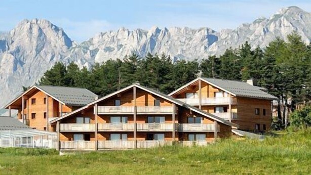 A mountain lodge with multiple wooden cabins, each with balconies, set against a backdrop of snow-capped mountains. - Madame Vacances Résidence La Crête du Berger (Le Dévoluy)