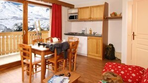 A cozy living room with a wooden dining table, chairs, a television, and a sofa. A kitchen with wooden cabinets and a window with a view of mountains is visible. - Madame Vacances Résidence La Crête du Berger (Le Dévoluy)