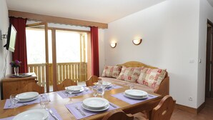 Ein Essbereich mit einem Holztisch, gedeckt für vier Personen, einer Couch mit gemusterten Kissen und Blick auf eine Holzterrasse durch eine Glastür.