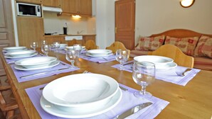Ein Esszimmertisch ist gedeckt mit Tellern, Besteck und Weingläsern. Im Hintergrund sind eine Küche und ein Sofa zu sehen.
