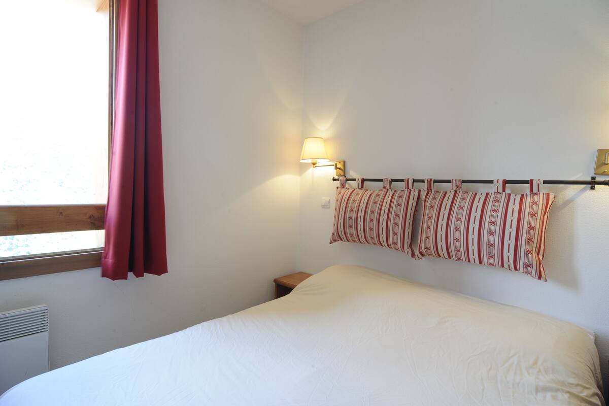 Un dormitorio con cama, mesita de noche, lámpara de pared y ventana con cortinas.