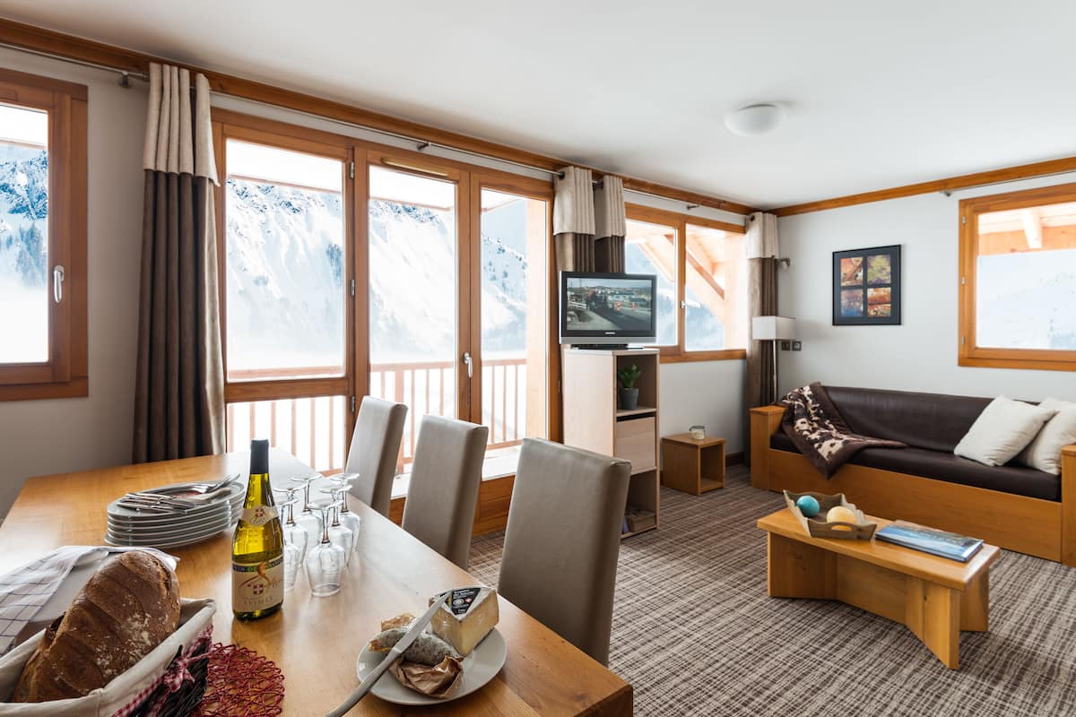 Une pièce chaleureuse avec une table à manger en bois, des chaises, un canapé, un téléviseur et une vue sur des montagnes enneigées, visible à travers de grandes fenêtres.