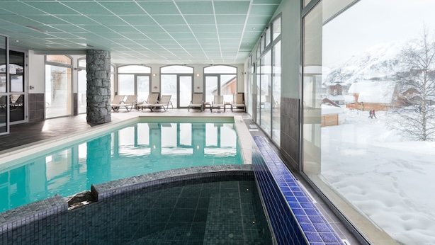 A swimming pool with a tiled edge, a stone pillar, and a view of snowy mountains through large windows. - Résidence Lagrange Vacances Les Balcons des Aiguilles (Fontcouverte-la-Toussuire)