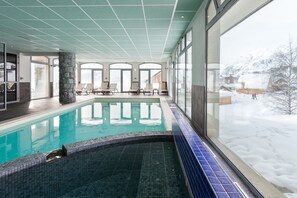 A swimming pool with a tiled edge, a stone pillar, and a view of snowy mountains through large windows. - Résidence Lagrange Vacances Les Balcons des Aiguilles (Fontcouverte-la-Toussuire)