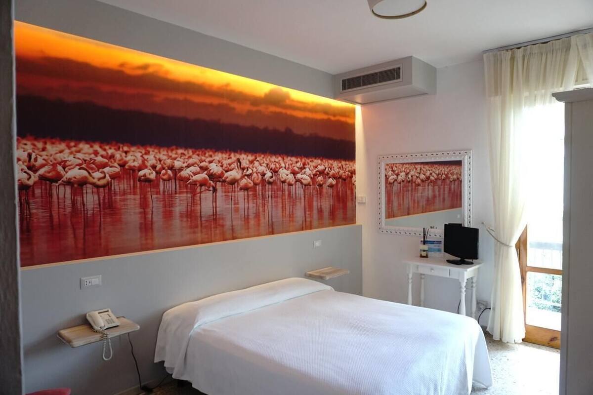 Ein Hotelzimmer mit einer großen Wandmalerei von Flamingos, einem Bett, einem Nachttisch mit Telefon, einem Fernseher und einem Fenster mit Vorhängen.