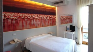 A hotel room with a large wall mural of flamingos, a bed, a bedside table with a phone, a TV, and a window with curtains. - Hotel Atlantico (Rosignano Marittimo)