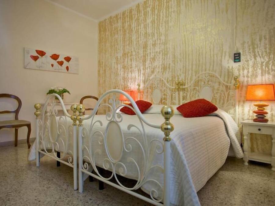 Een slaapkamer met een bed met wit metalen frame, een nachtkastje met lamp, een stoel en een muur met een structuurbehang en een bloemschilderij.