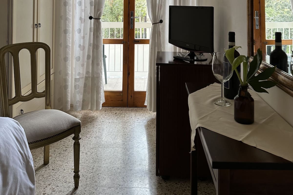 Ein Zimmer mit Holzstuhl, einem Tisch mit Weinglas und Flasche, einem Fernseher und einem Balkon mit Vorhängen.