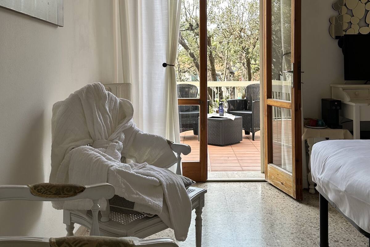 Ein Schlafzimmer mit einem Bett, einem Sessel mit Bademantel, einem Tisch mit Tastatur und Blick auf eine Außenpatio mit Möbeln und Bäumen.