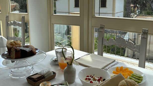 A dining table with a variety of food items, a vase with a flower, and a view of trees and buildings through large windows. - Hotel Atlantico (Rosignano Marittimo)