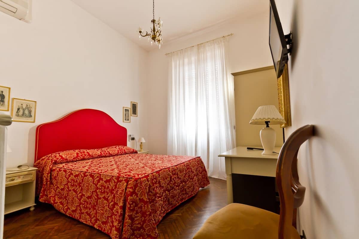 A room with a red bed, a wooden chair, a desk with a lamp, and a mirror on the wall.