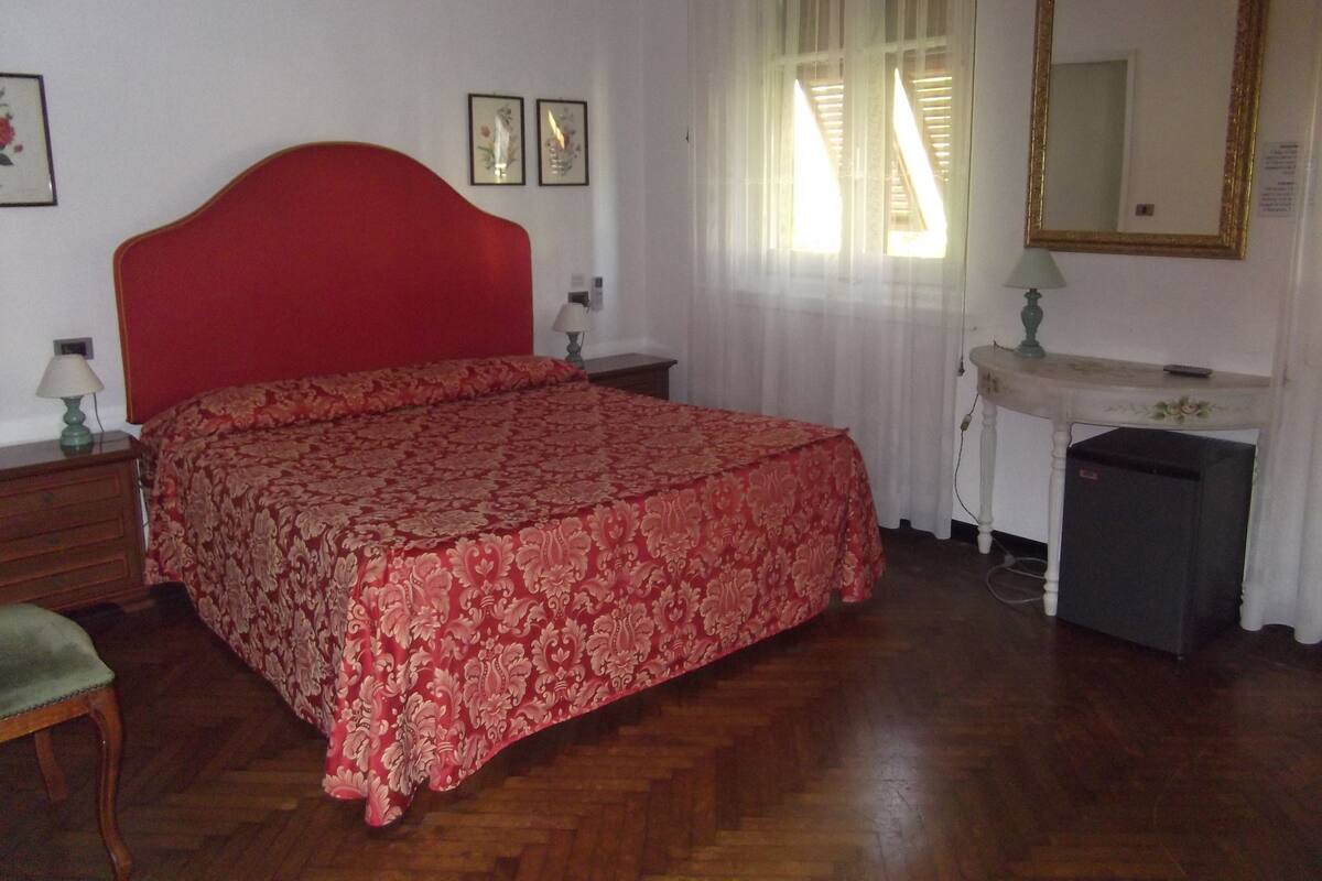 A bedroom with a red bedspread, a wooden dresser, a mirror, and a window with curtains.