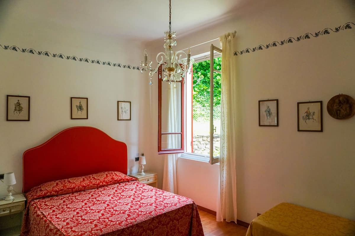 A bedroom with a red headboard, a bed with a red and white patterned bedspread, a bedside table with a lamp, a window with curtains, and framed pictures on the wall.