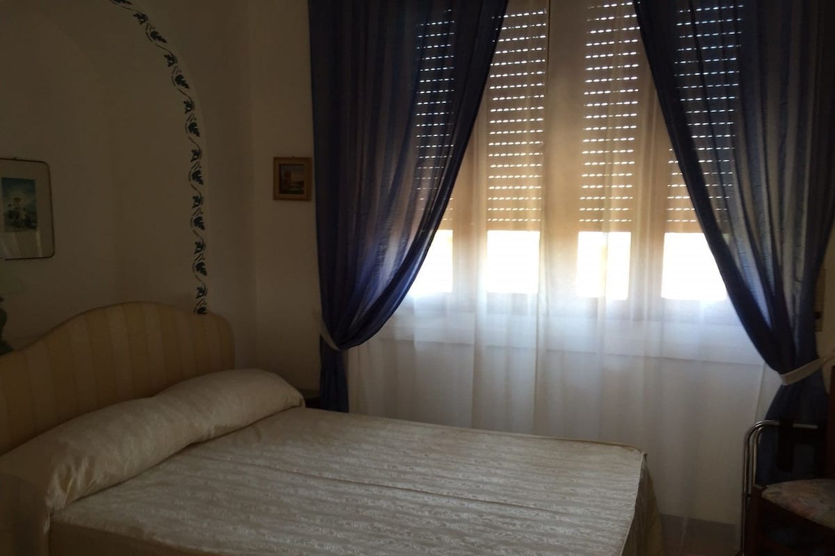 A bedroom with a bed, curtains, a chair, and a wall with a picture.