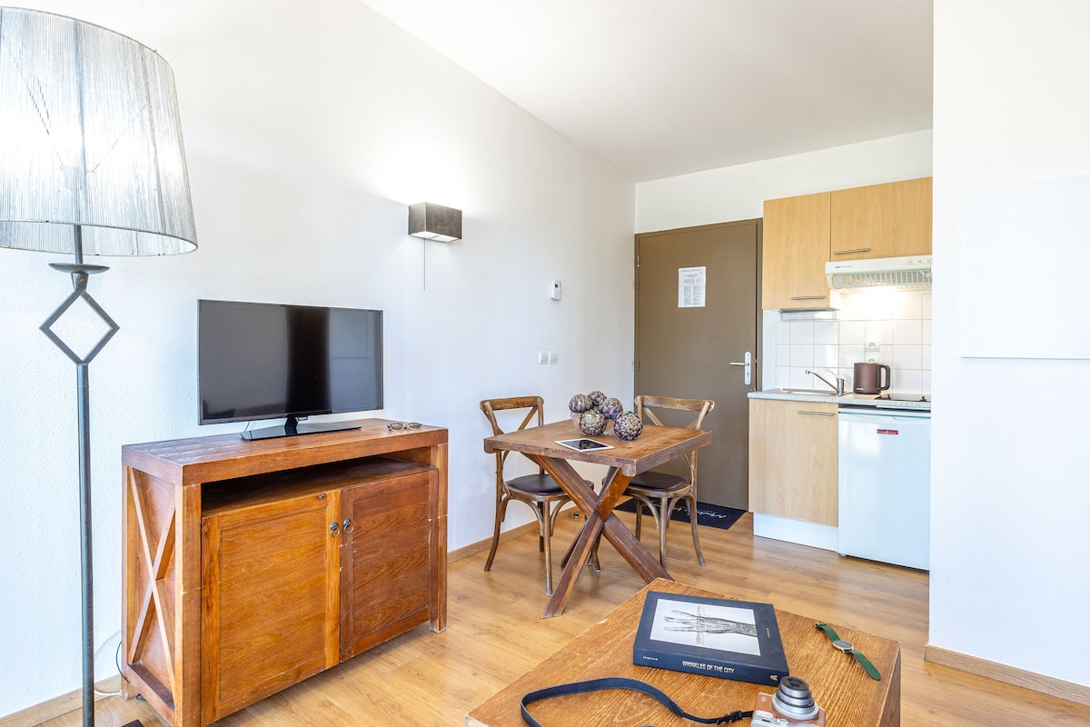 Un espace de vie compact comprenant un meuble de rangement en bois servant d’unité multimédia, un téléviseur à écran plat, une table à manger avec des chaises, un coin cuisine avec un réfrigérateur et une lampe.