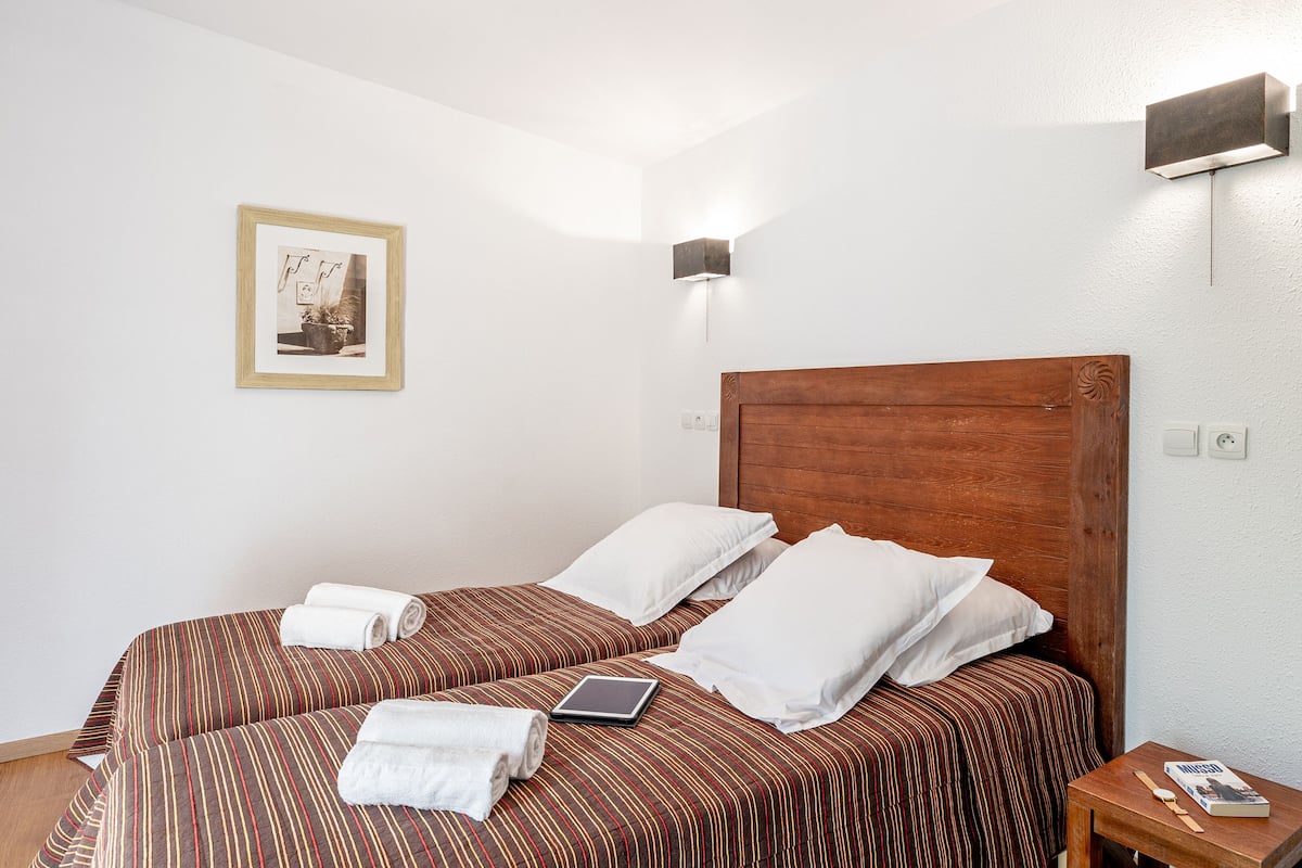 Une chambre d’hôtel avec deux lits, une table de chevet avec un livre, une lampe fixée au mur et un tableau encadré accroché au mur.