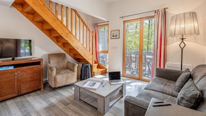 A cozy living room with a wooden staircase, a sofa, a coffee table with a laptop, and a TV on a wooden cabinet.