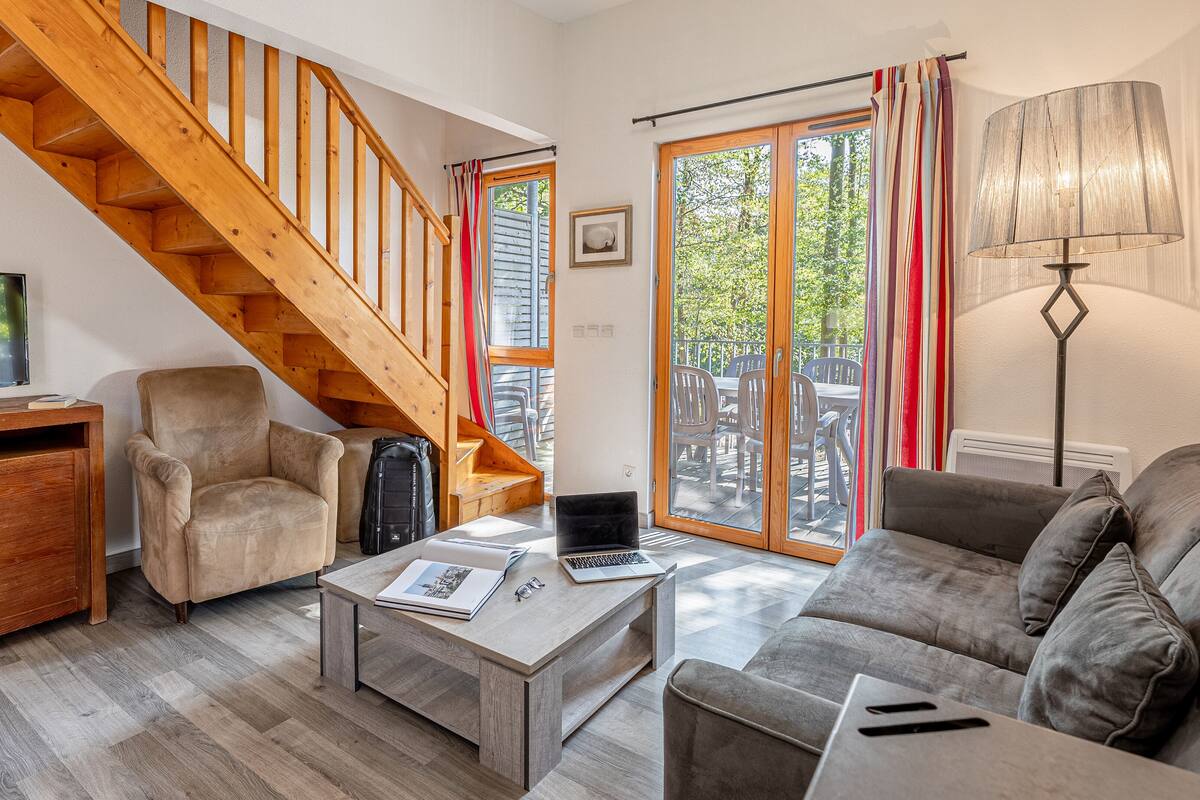Un salon chaleureux avec un escalier en bois, un canapé, une table basse sur laquelle se trouve un ordinateur portable, et un téléviseur posé sur une commode en bois.