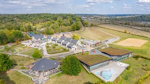 An aerial view of a modern building complex with multiple structures, including a large glass building with a swimming pool, surrounded by green spaces and a parking area.