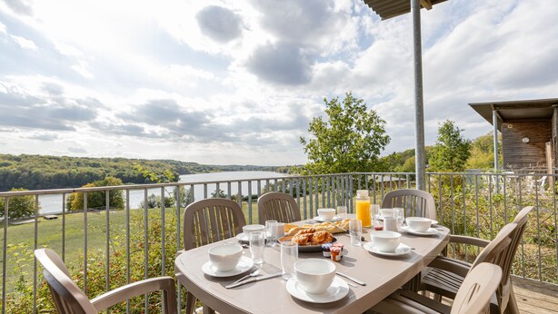 A table set for breakfast with a scenic lake view, outdoor chairs, and a wooden deck.