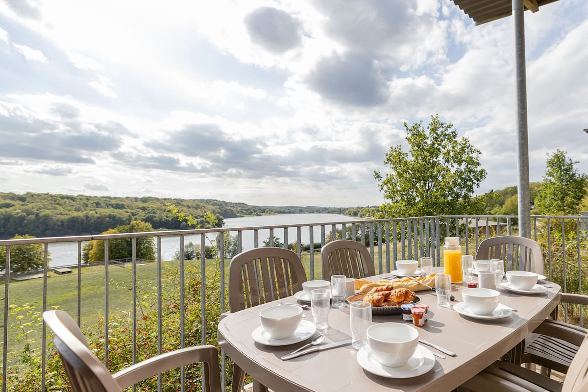 Une table dressée pour le petit-déjeuner, avec une vue pittoresque sur le lac, des chaises d’extérieur et une terrasse en bois.