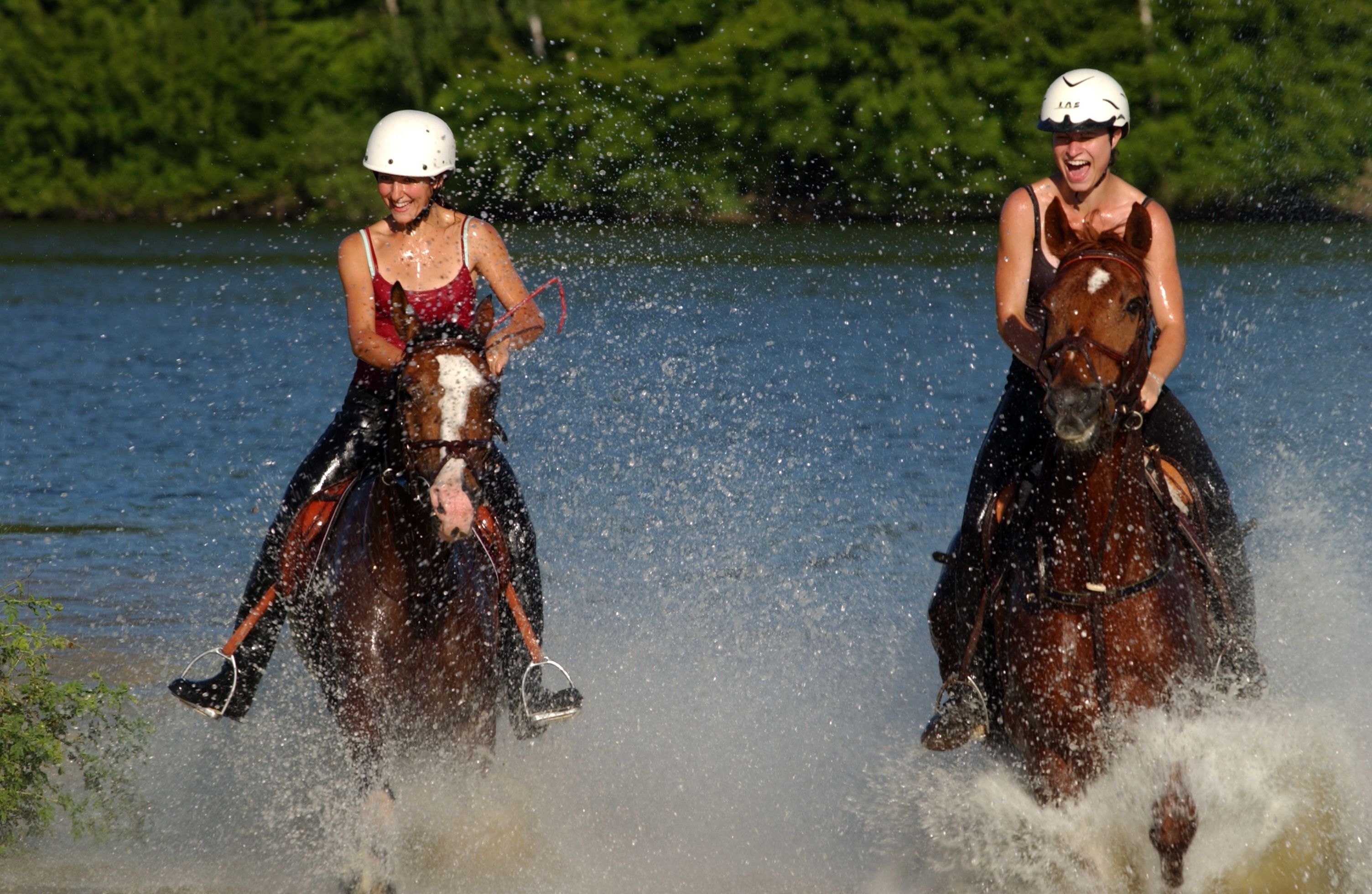 Duas pessoas cavalgam a cavalo por um corpo de água, espalhando água.
