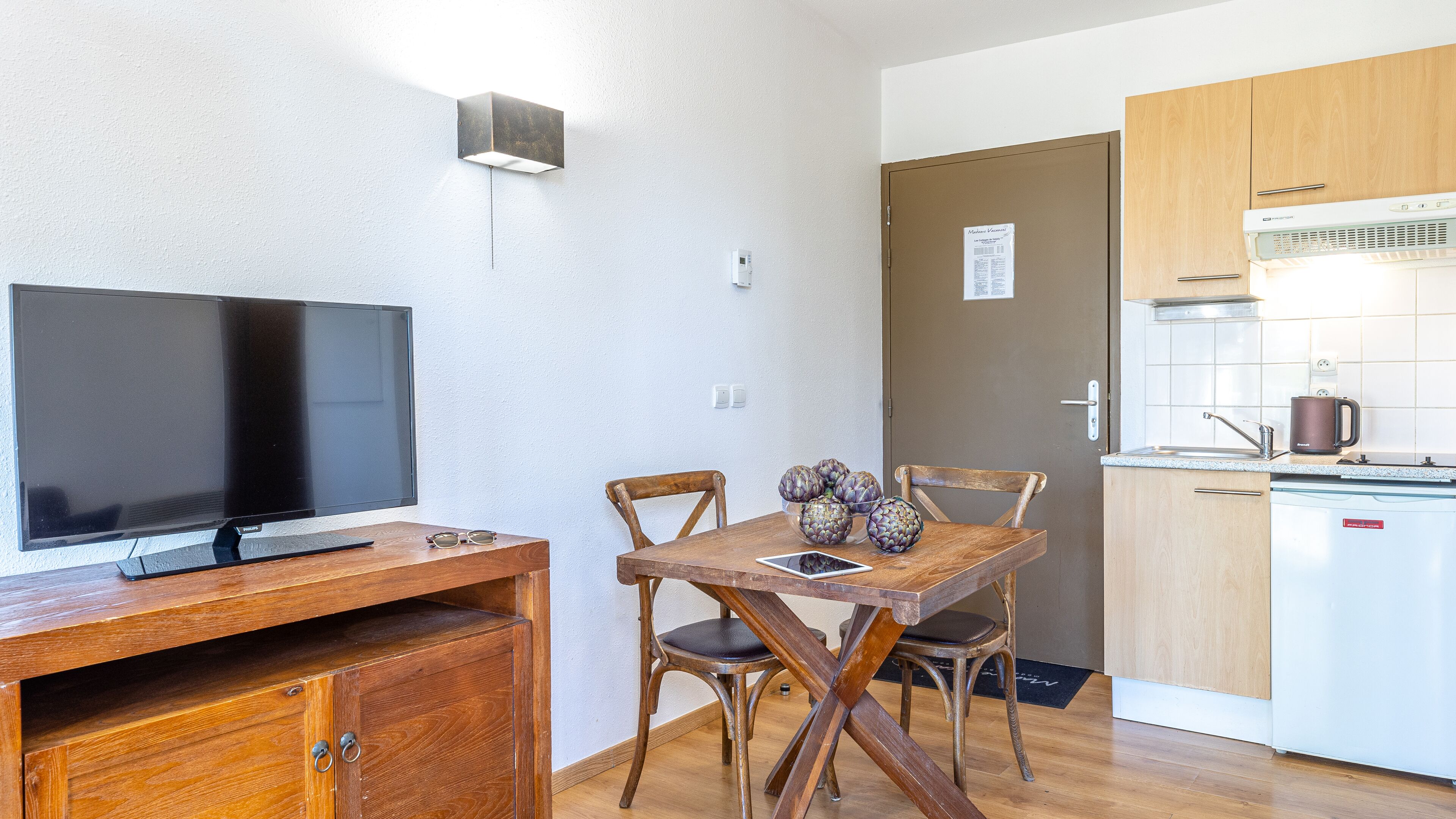 Cozinha compacta com fogão embutido, área de refeições com mesa e cadeiras de madeira, e uma TV de tela plana sobre um armário de madeira.