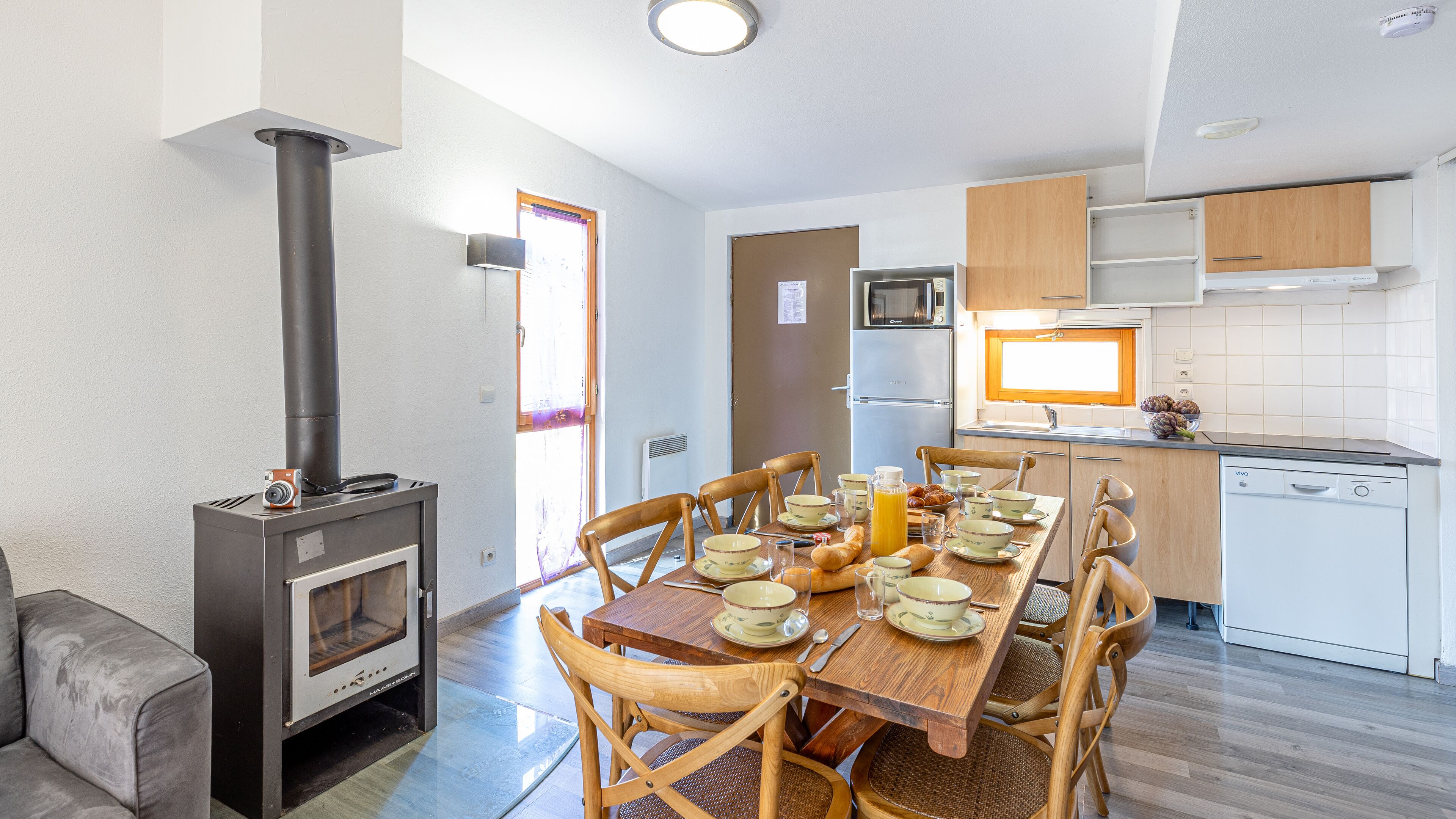 A compact kitchen with a wood-burning stove, a dining table set for four, and a compact refrigerator.