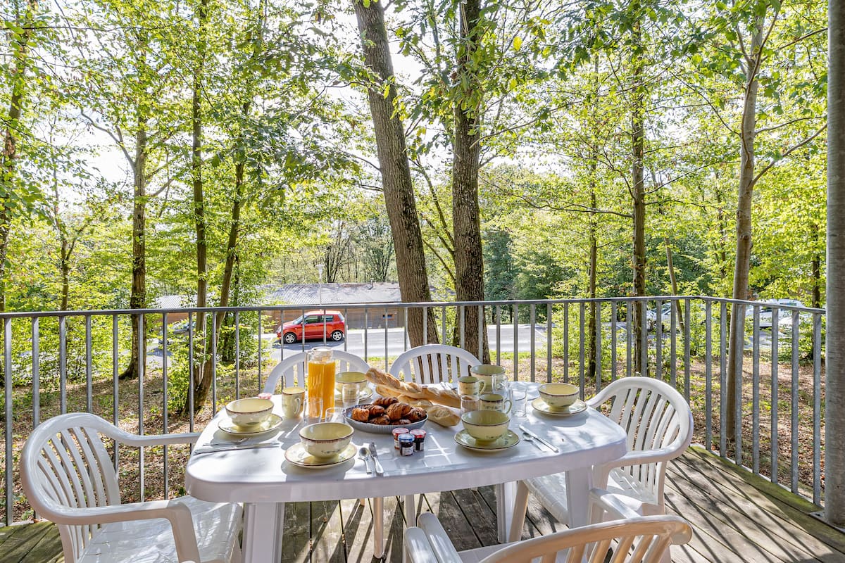 Une table dressée à l’extérieur, entourée de chaises blanches et d’une rambarde en métal, offre une vue sur une zone boisée avec des arbres et une voiture rouge, visible au loin, sur une route.